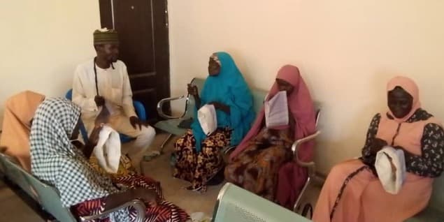 A facilitator speaking with a semicircle of seated women in a community hall, each holding a white cloth item.