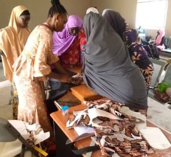 Women gathered around a workbench cutting and assembling patterned fabric pieces during a practical skills session.