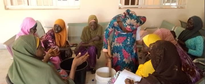 A circle of women seated around containers and a pestle, one standing and demonstrating to the group.